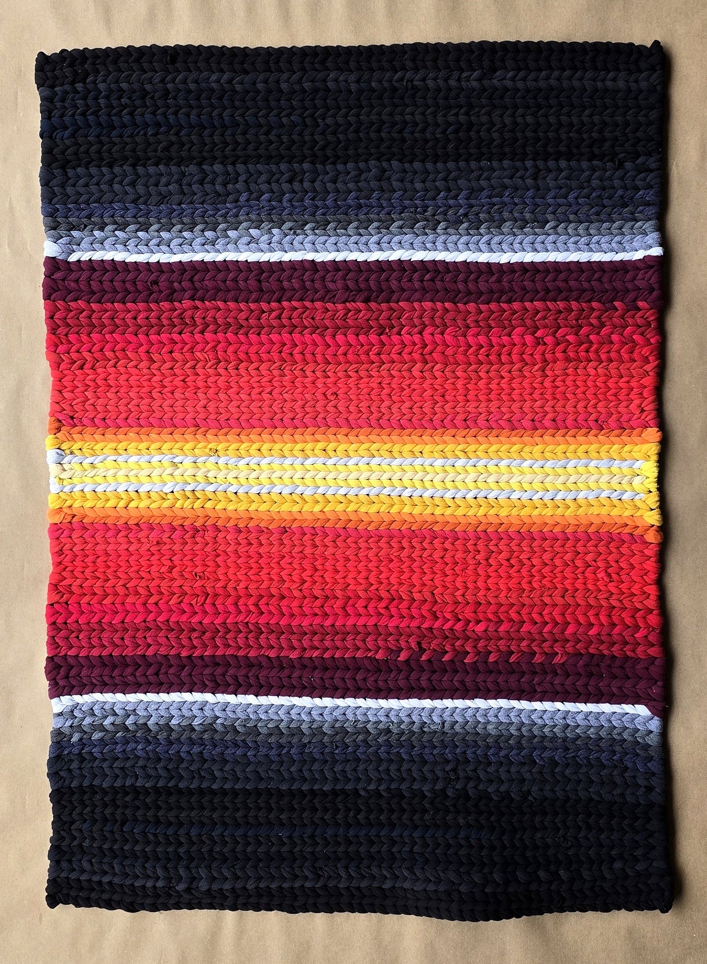 A hand-twined rectangular rug with deep charcoal edges and a glowing center band of red, orange, and gold, inspired by ember light and crafted from repurposed T-shirt knits.