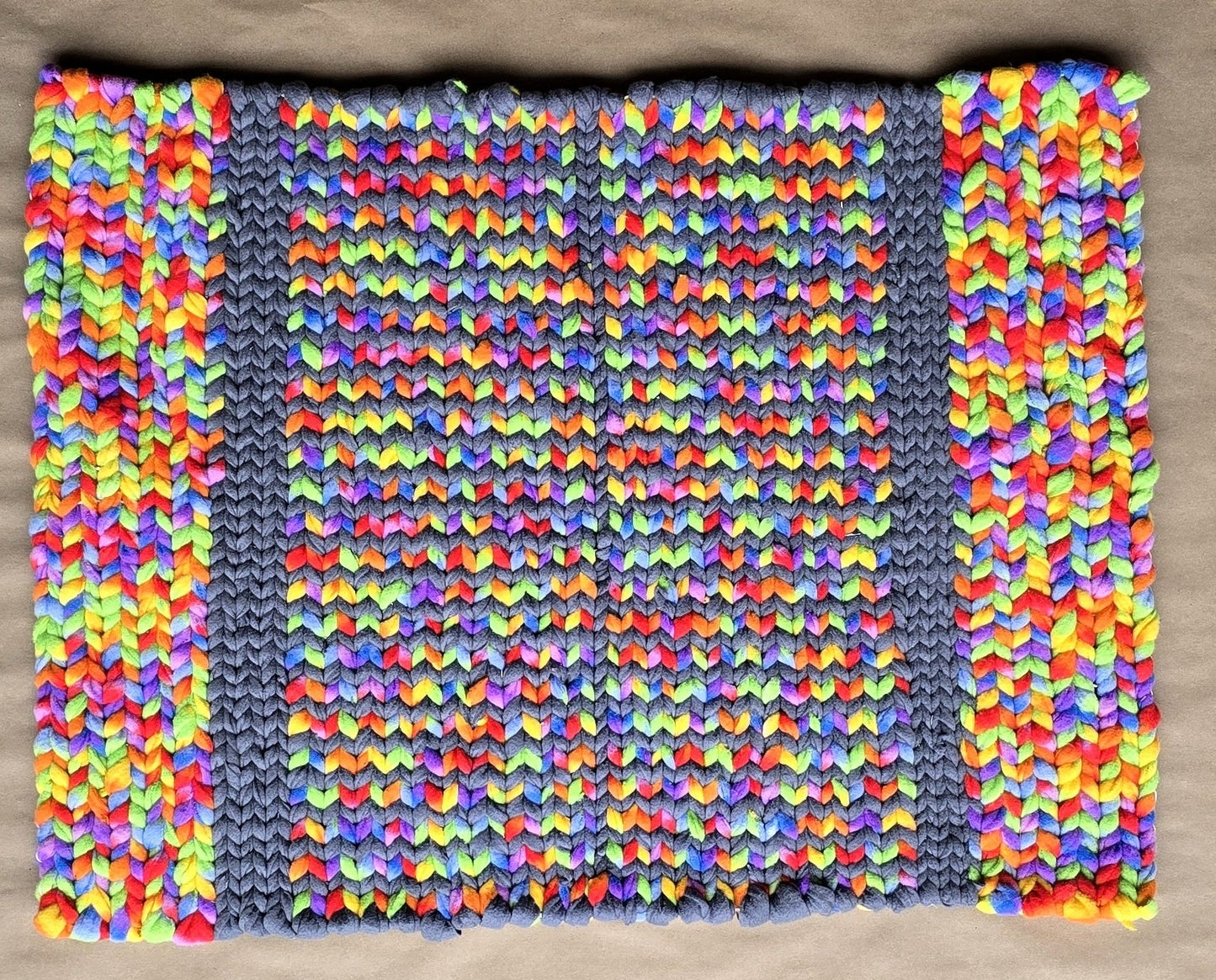 A hand-twined rectangular rug made from repurposed fleece, featuring bright multicolor strands woven in a repeating pattern between gray vertical bands, displayed on kraft paper.