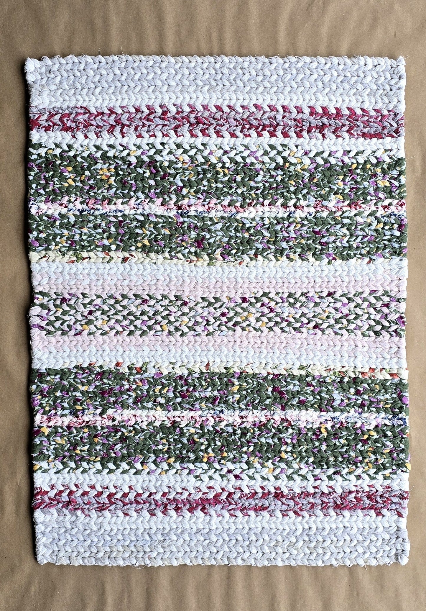 A hand-twined rectangular flannel rug with repeating horizontal stripes of frosted white, evergreen, and muted berry tones, displayed on kraft paper.