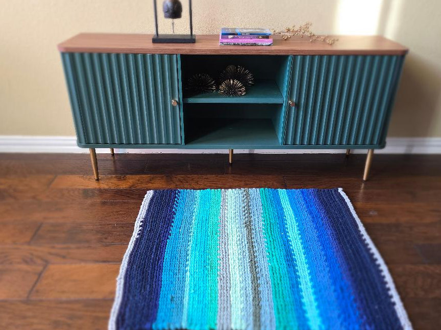 Handwoven blue and teal striped rug styled beneath a green console table in a calm, modern living space.