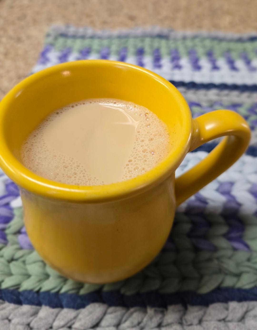 A yellow ceramic mug filled with warm spiced oat milk, resting on a woven mat on a kitchen counter.