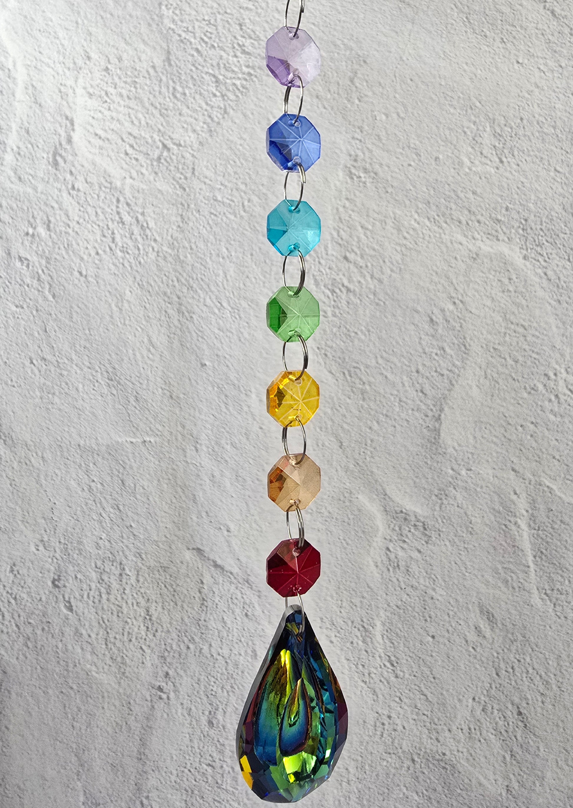 Sevenfold Halo crystal suncatcher with a vertical rainbow spectrum of faceted glass crystals and a prismatic aurora-style teardrop pendant, hanging against a light plaster background.