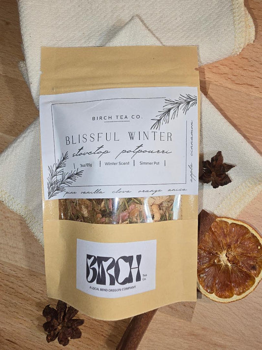 Kraft pouch of Blissful Winter simmer pot potpourri displayed on folded cream towels with dried orange slices and star anise, showing the illustrated front label.