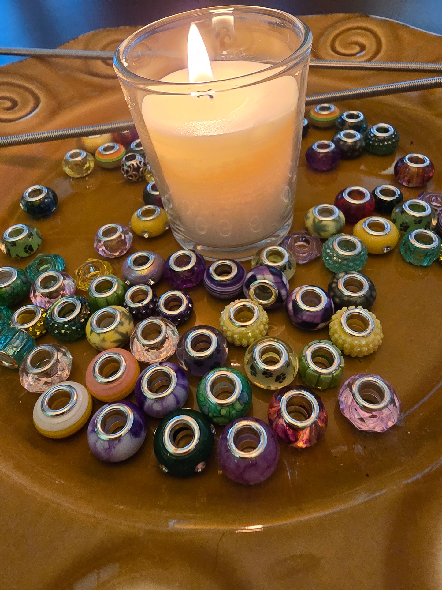 Assorted glass and acrylic beads arranged around a lit candle on a warm-toned ceramic plate with metal garden rods in the background.