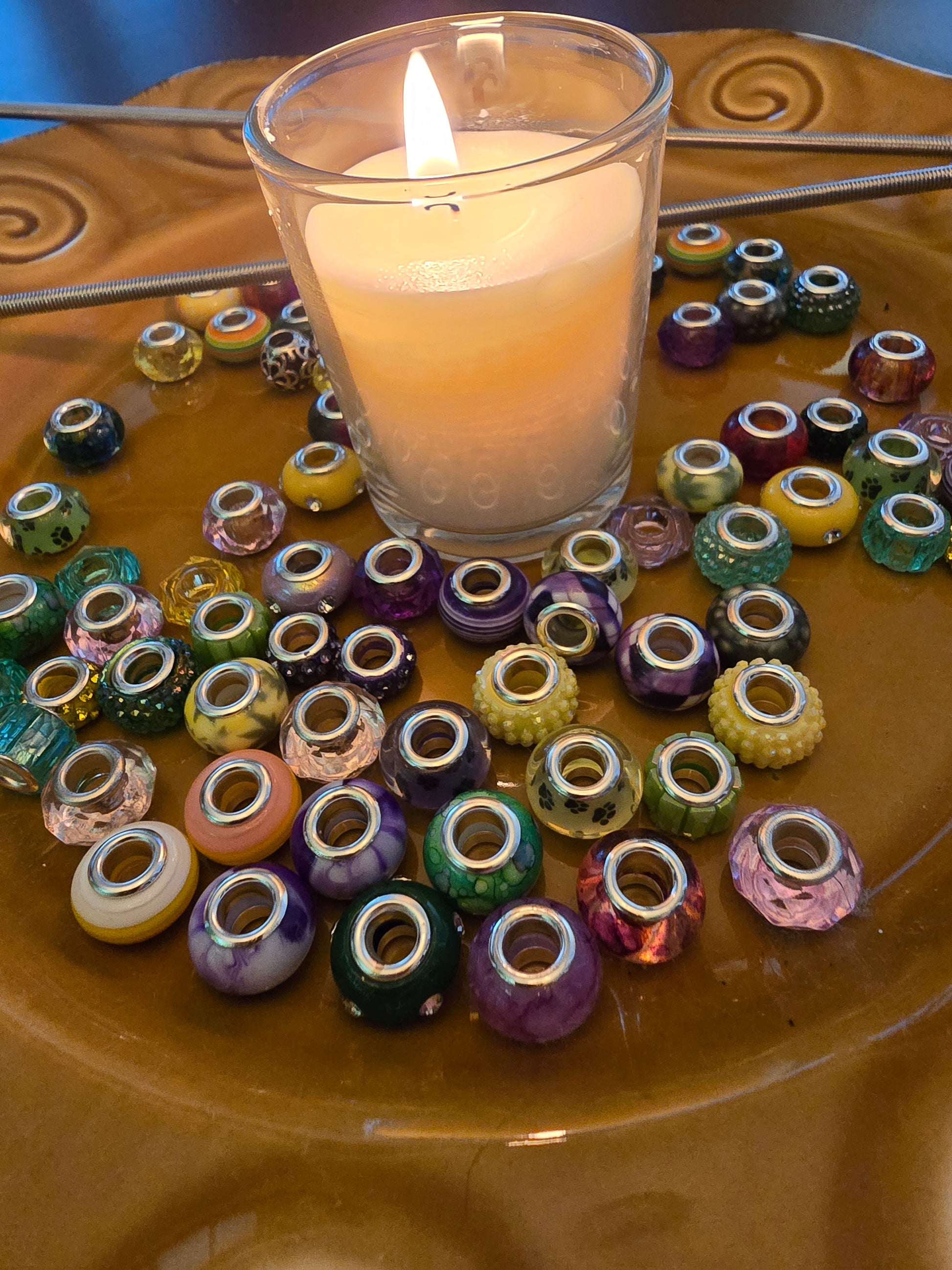 Assorted glass and acrylic beads arranged around a lit candle on a warm-toned ceramic plate with metal garden rods in the background.