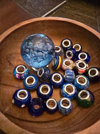 Blue-toned glass and acrylic beads with metal cores arranged in a wooden bowl beside a decorative glass sphere.