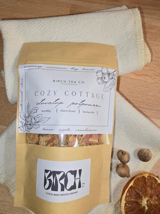 Kraft pouch of Cozy Cottage simmer pot potpourri displayed on a folded cream towel with dried orange slices and nuts, showing the illustrated front label.