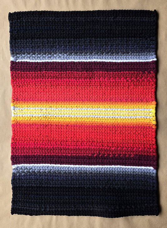 A hand-twined rectangular rug with deep charcoal edges and a glowing center band of red, orange, and gold, inspired by ember light and crafted from repurposed T-shirt knits.