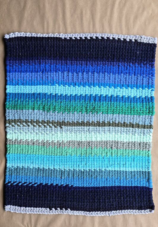 A handwoven rectangular rug featuring horizontal stripes in deep navy, royal blue, aqua, teal, and soft gray tones, displayed on a kraft-paper background.
