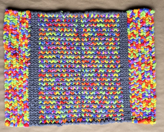 A hand-twined rectangular rug made from repurposed fleece, featuring bright multicolor strands woven in a repeating pattern between gray vertical bands, displayed on kraft paper.