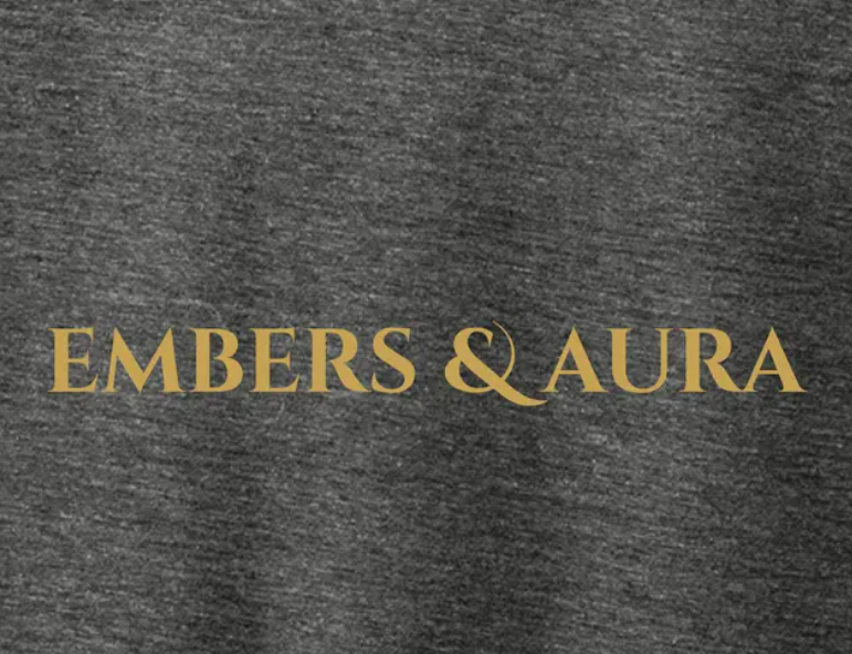 Close-up of the Embers & Aura gold logo printed on the front of a gray tri-blend T-shirt.