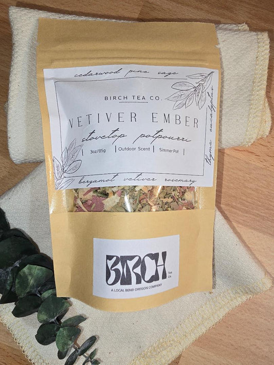 A kraft paper pouch of Vetiver Ember simmer pot potpourri displayed on a folded cream towel with eucalyptus leaves beside it. The label shows botanical illustrations and the scent name, with dried botanicals visible through the window at the bottom