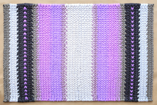 Hand-twined rectangular rug in black, gray, and layered violet tones, with subtle heart-shaped accents woven into the design.