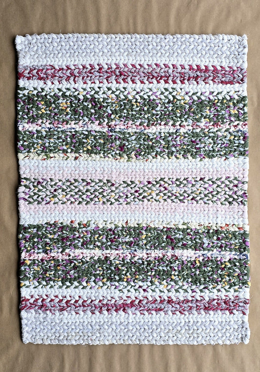 A hand-twined rectangular flannel rug with repeating horizontal stripes of frosted white, evergreen, and muted berry tones, displayed on kraft paper.