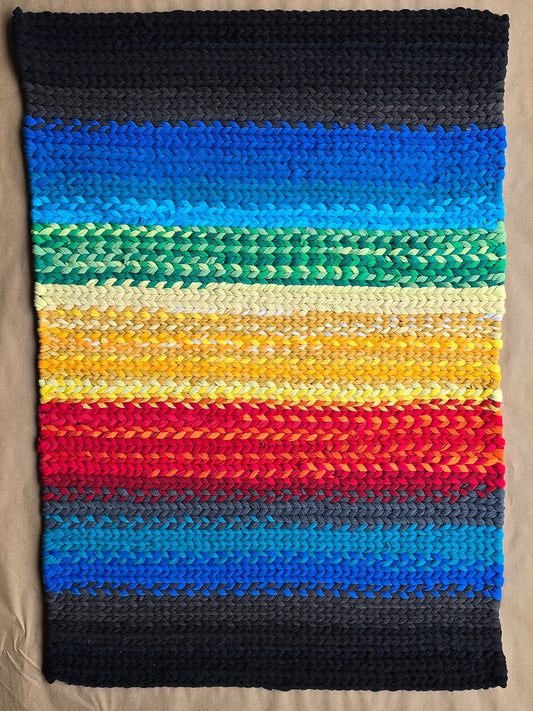 A hand-twined rectangular rug made from repurposed T-shirt knits, featuring horizontal bands of deep navy, bright blues, greens, yellows, reds, and oranges, displayed on kraft paper.
